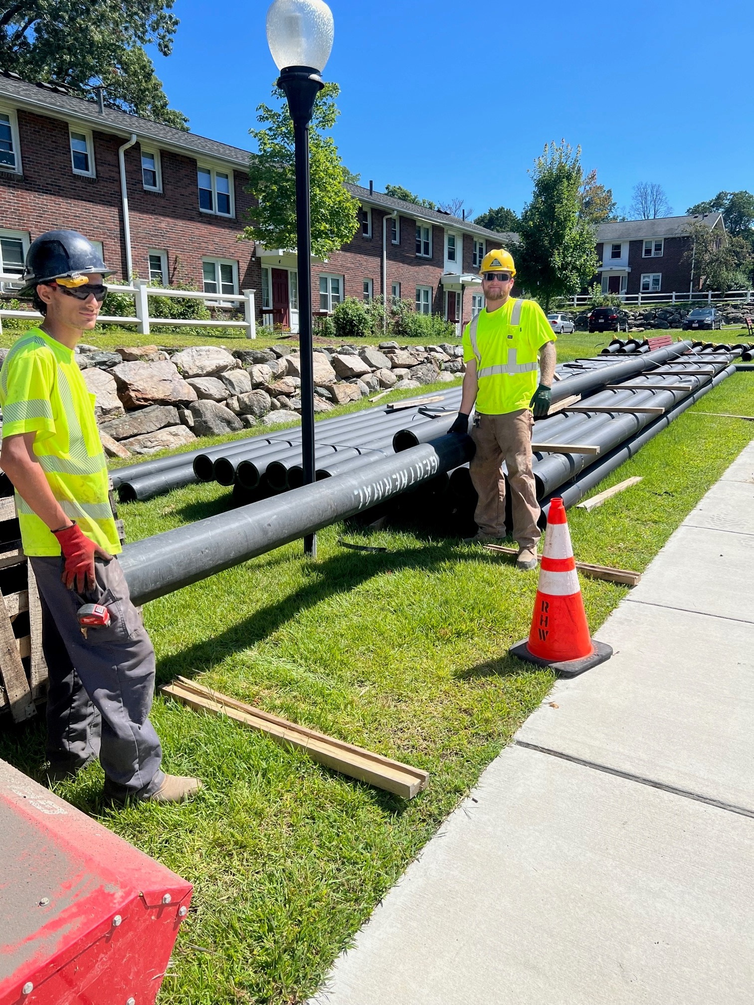 Two workers in hard hats stand by pipe on a sunny day.