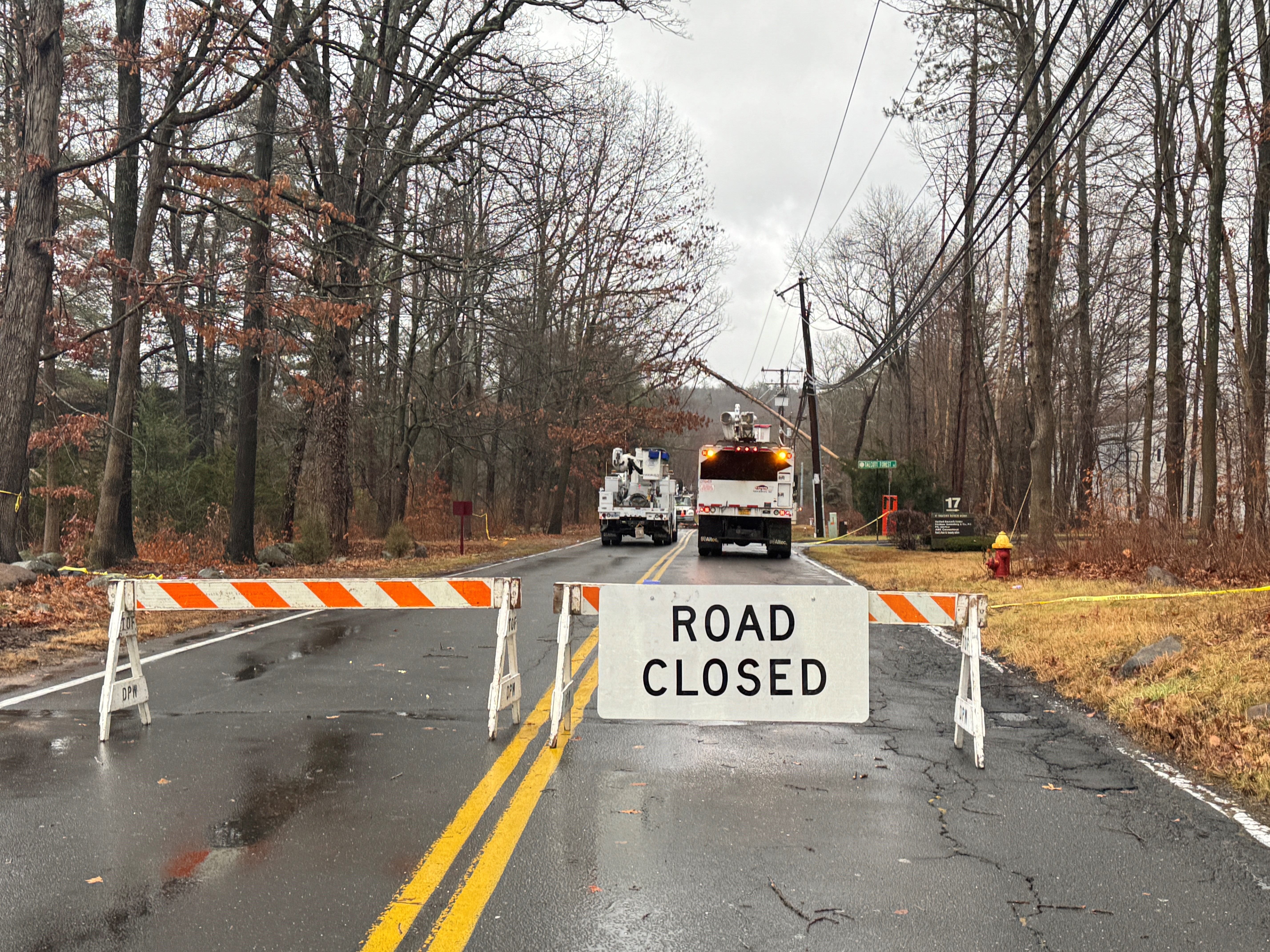 Barricade with “ROAD CLOSED” sign blocks a wet two-lane road. Two utility trucks with flashing lights are parked ahead near downed power lines. Bare trees line both sides under a gray, overcast sky.