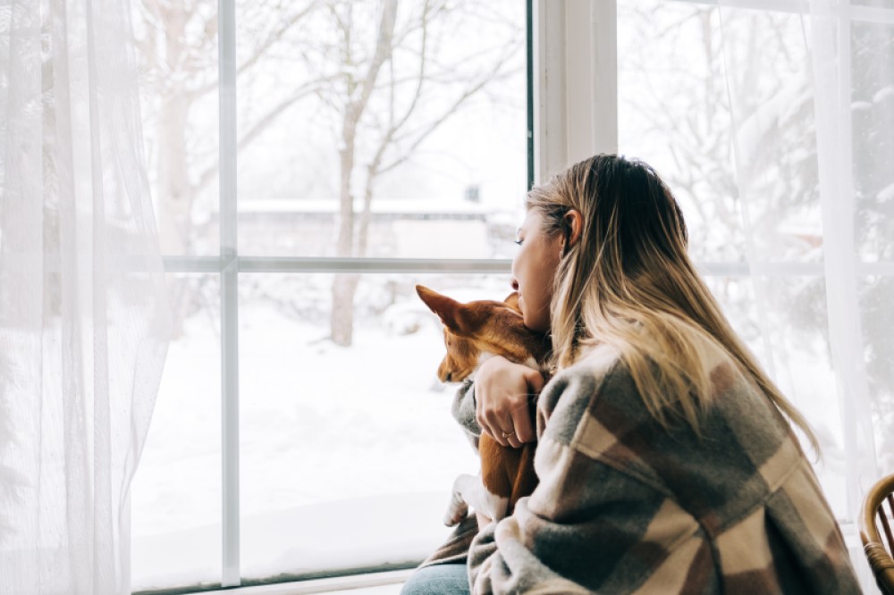 A woman holds a dog and looks out her window into a snowy yard