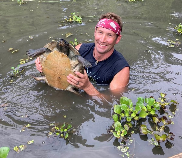 Artist Matt Patterson standing in chest-deep water, smiling while holding a large turtle.